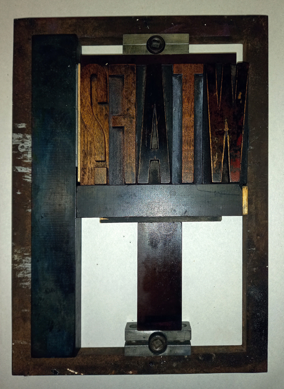 The photo shows five antique wooden typeface blocks that spell out the acronym W T A F and a question mark. The letters are set back to front in a metal frame, a chase, and various pieces of metal furniture and a couple of groynes have been used to hold the letters firm ready for printing.